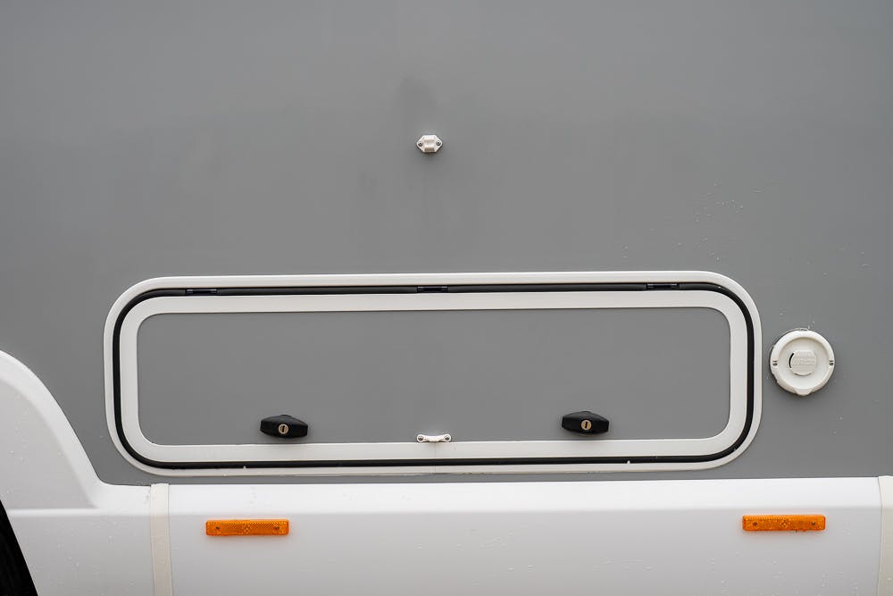 A close-up of the side of a 2022 Bailey Autograph 74-4 motorhome. The image showcases a rectangular storage compartment with a white frame, secured by two black latches. The side of the motorhome is primarily gray with a white bottom and features orange reflectors near the base.