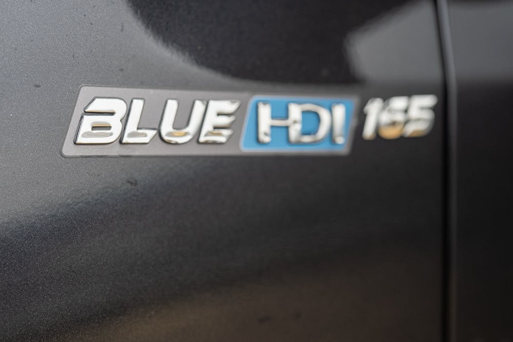 Close-up of a 2022 Bailey Autograph 74-4 vehicle's exterior displaying a badge with the text "BLUE HDi 165" in metallic letters, where "HDI" is highlighted in blue.