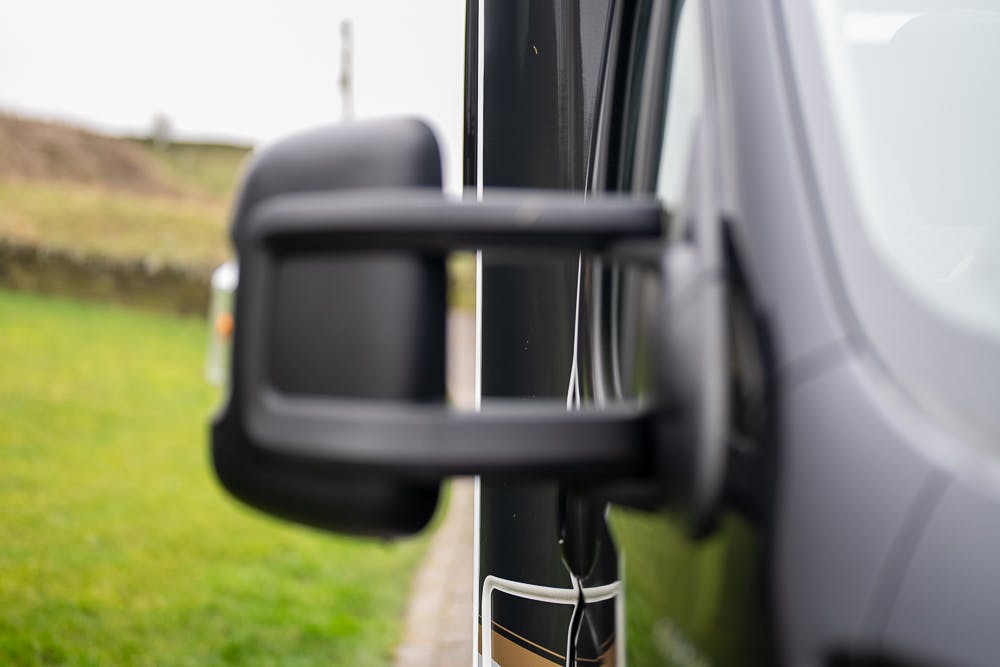 A close-up view of the side mirror of a black 2022 Bailey Autograph 74-4, with the door and part of the window visible. The background shows a blurred landscape with a grassy area and partial view of a stone wall.