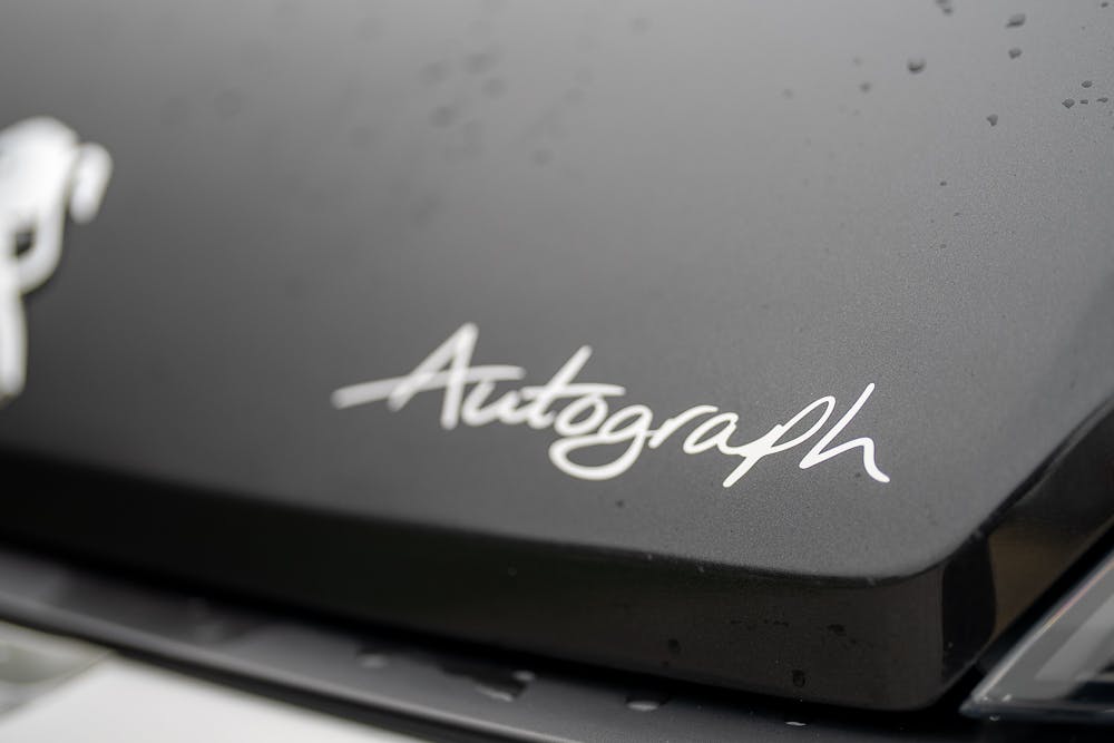 Close-up image of the word "Autograph" written in a cursive font on what appears to be a part of a 2022 Bailey Autograph 74-4 vehicle. The surface has water droplets, suggesting recent contact with moisture, possibly rain.