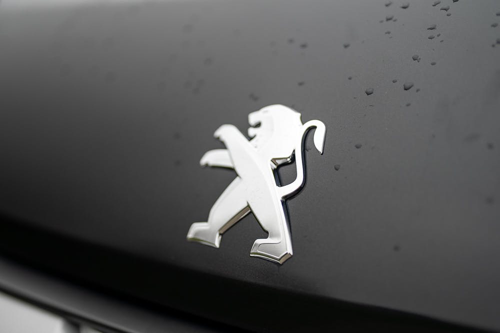 Close-up shot of a silver Peugeot lion emblem on a matte black surface with water droplets, capturing the refined elegance reminiscent of the 2022 Bailey Autograph 74-4.