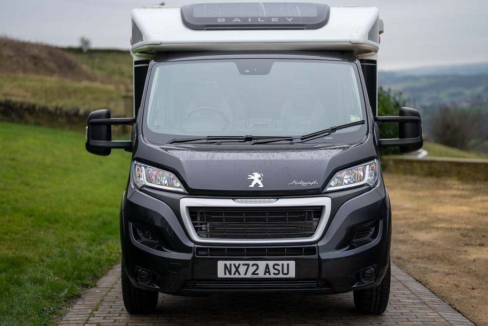 A recreational vehicle with a "Bailey" logo on the front is photographed head-on. The 2022 Bailey Autograph 74-4, adorned with a Peugeot emblem, displays a license plate reading "NX72 ASU." It is parked on a path bordered by grassy areas and distant countryside scenery.