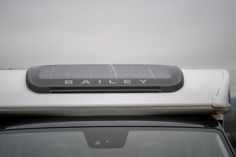 Close-up of the roof section of a 2022 Bailey Autograph 74-4 motorhome. The word "Bailey" is prominently displayed on the grey panel near the top of the vehicle. The panel appears damp, likely due to recent rain, against a cloudy sky background.