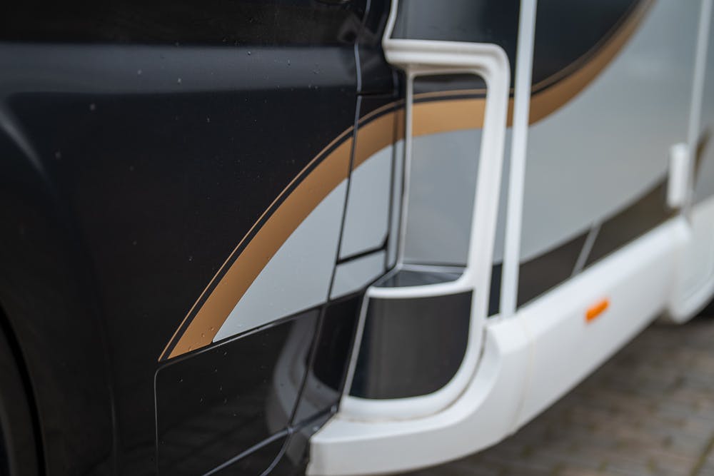 Close-up of the side of a black-and-white 2022 Bailey Autograph 74-4 vehicle with a gold and gray stripe design. The image shows a portion of the vehicle's exterior, including a white structural component and a small orange reflector. The background is slightly blurred pavement.