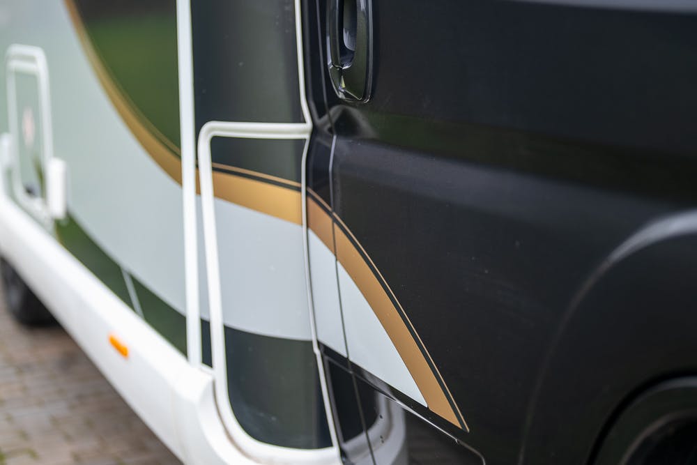 Close-up view of a black 2022 Bailey Autograph 74-4 with white and brown design accents on its side. The image prominently features the vehicle's door and part of a white structural frame. The ground beneath the vehicle is paved with brick.