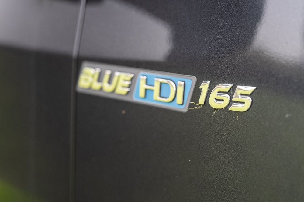 Close-up of a car badge displaying the text "BLUE HDI 165" on a black vehicle. The badge is situated near a scratch on the car's surface, reminiscent of the meticulous detailing seen in models like the 2022 Bailey Autograph 74-4.