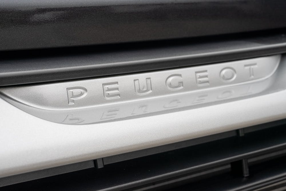 Close-up of a silver Peugeot car emblem on the front grille of the 2022 Bailey Autograph 74-4. The emblem, engraved with "Peugeot" in uppercase letters, sits prominently in the center of the grille against a backdrop of dark and light gray tones.