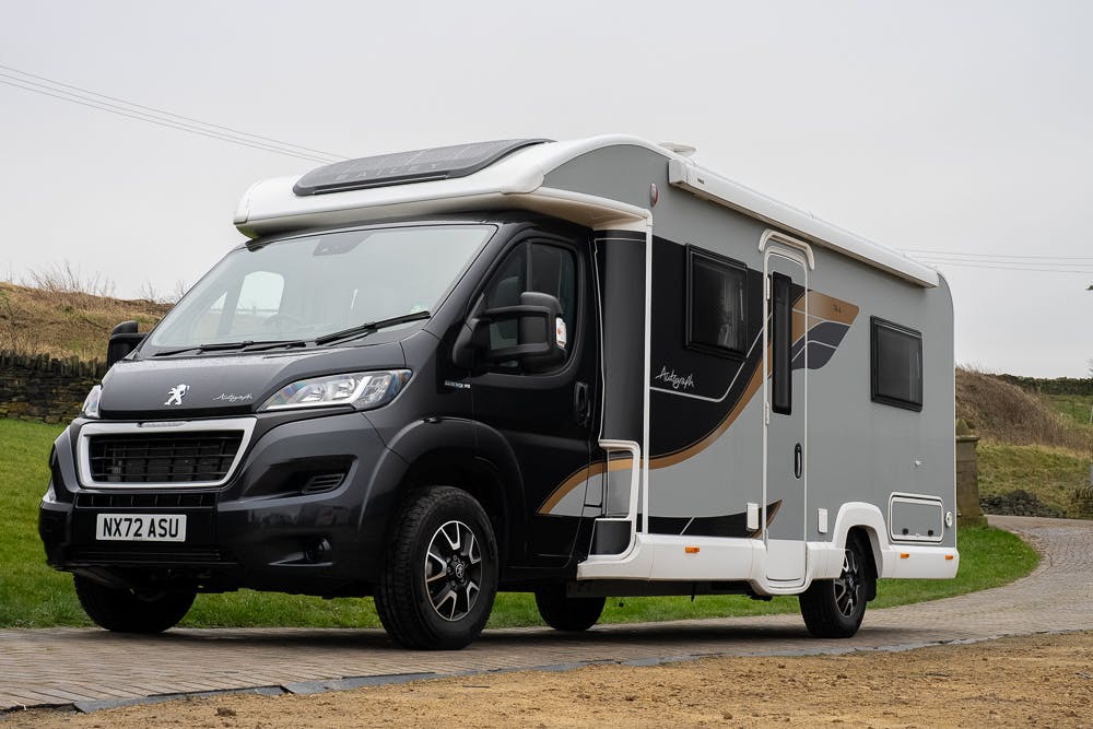 A black and gray 2022 Bailey Autograph 74-4 motorhome with a UK license plate "NX72 ASU" is parked on a paved surface. The vehicle features a large side window, a solar panel on the roof, and modern design elements. A grassy hill and stone wall are visible in the background.