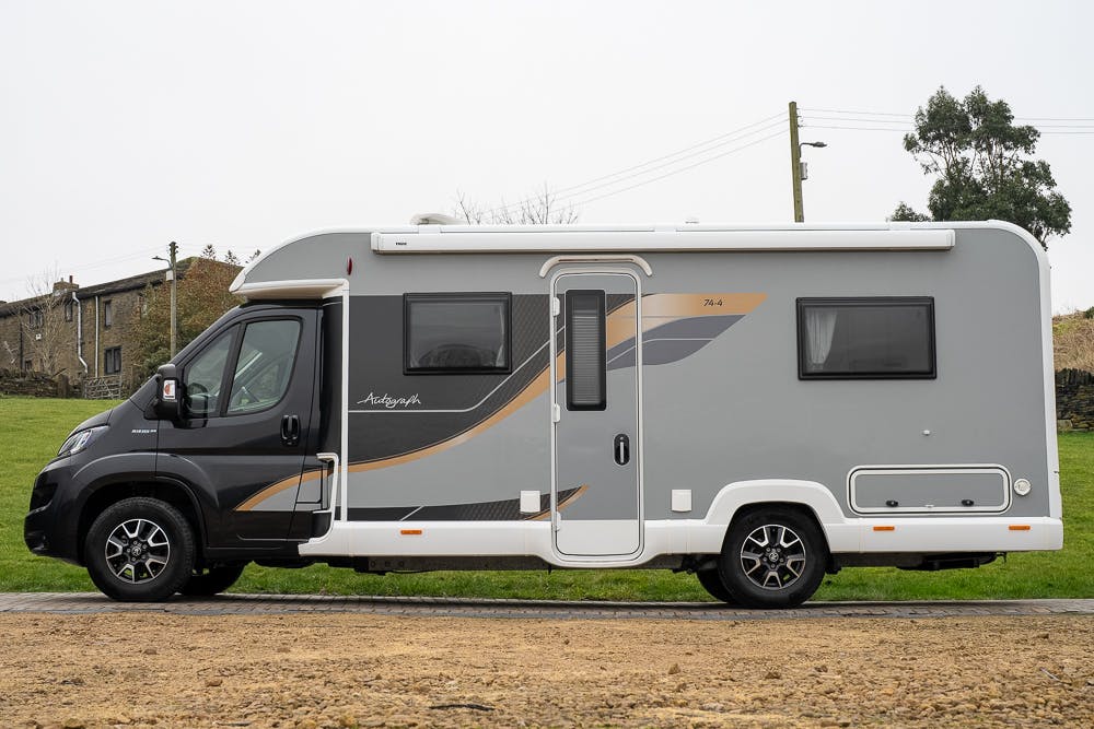 A side view of the 2022 Bailey Autograph 74-4, a modern gray motorhome with black and gold accents, parked on a gravel surface with a grass area and a brick building in the background. The motorhome features several windows and proudly displays the brand name "Autograph" on the side.