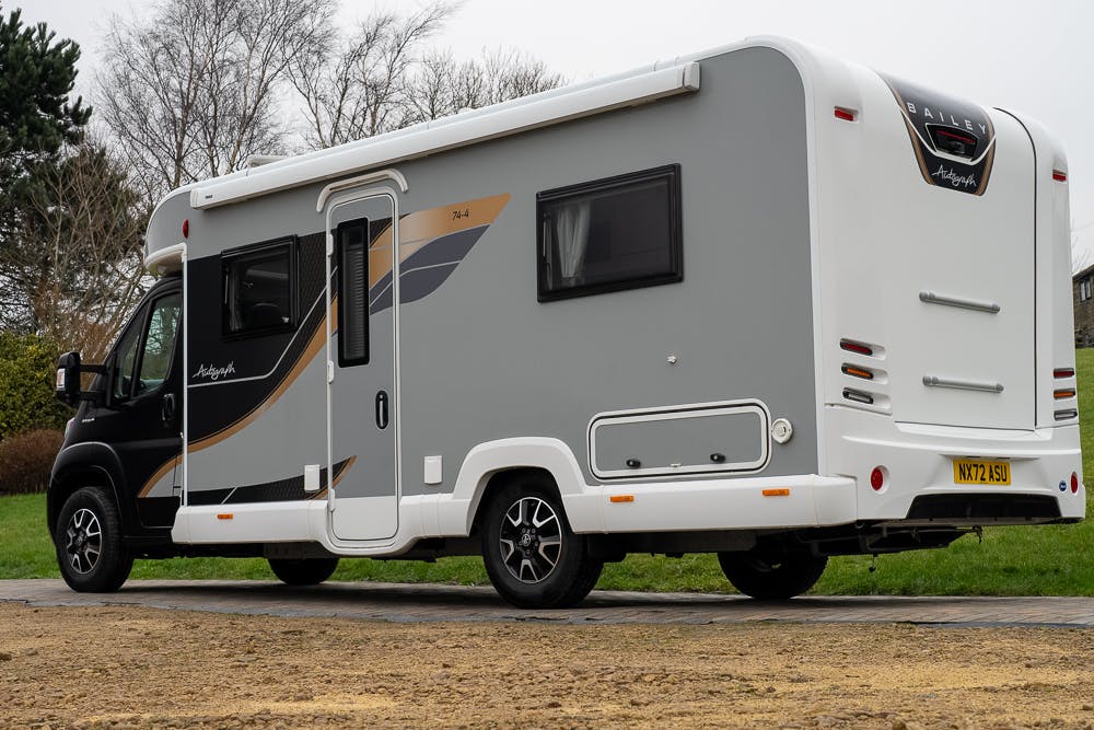 A gray and white 2022 Bailey Autograph 74-4 motorhome is parked outdoors on a paved area with a grassy surface nearby. The vehicle has tinted windows, a side door, and various compartments. The registration plate reads "NX71 YSL." Trees and bushes are visible in the background.