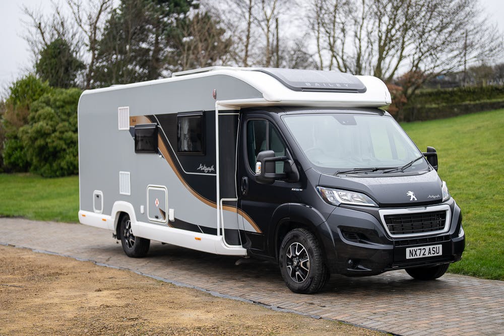 A grey, modern 2022 Bailey Autograph 74-4 motorhome with a black front is parked on a paved driveway. The vehicle has multiple windows and a sleek design. Grass and trees are visible in the background. The motorhome has a license plate reading "NX72 ASU.