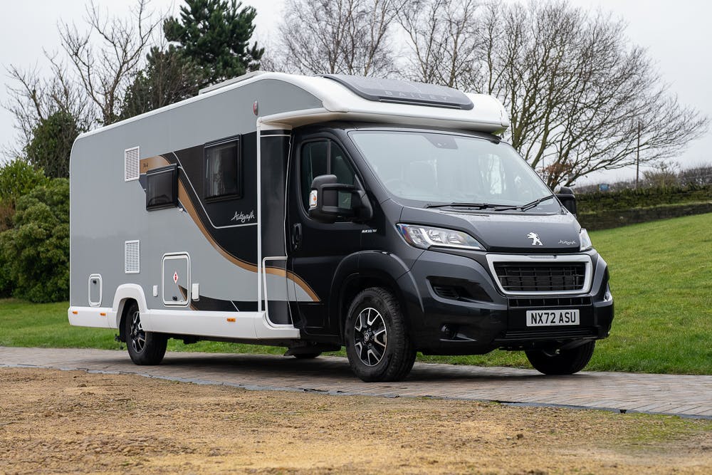 A modern 2022 Bailey Autograph 74-4 motorhome is parked on a paved area. The vehicle is gray with black and gold accents, featuring a Peugeot logo on the front. The license plate reads NX72 ASU. Trees and a grassy area are visible in the background on an overcast day.