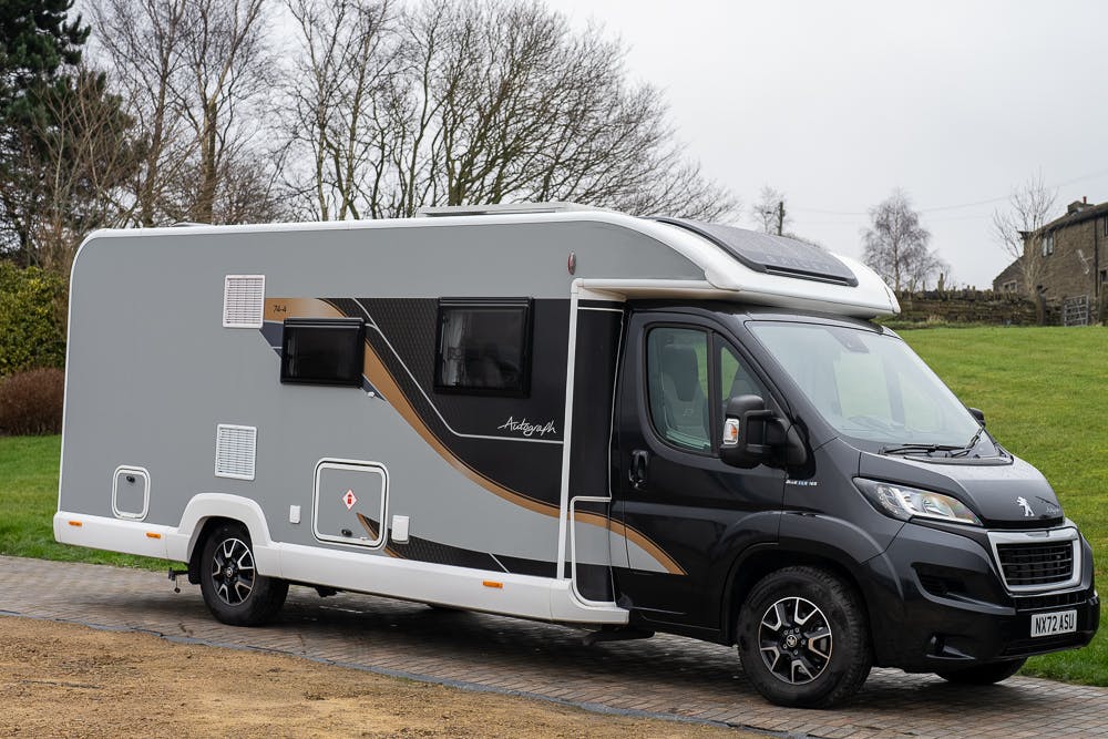 A gray and white 2022 Bailey Autograph 74-4 motorhome is parked on a brick driveway with grass and trees in the background. The vehicle features a white roof, black wheels, and a side window. The word "Autograph" adorns the side, and its license plate reads "N17X ASL.