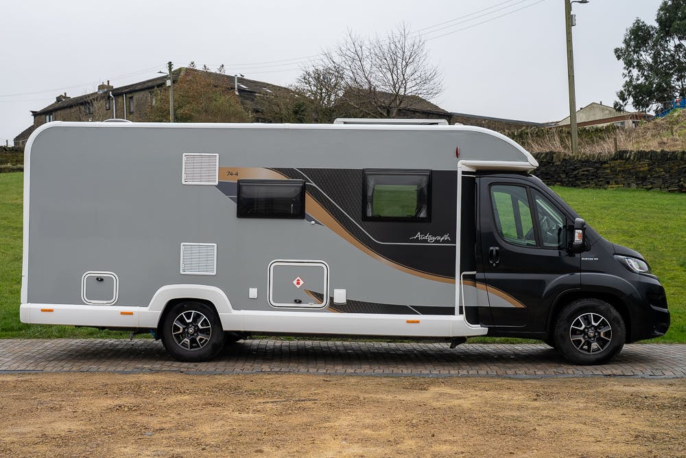 A gray and black 2022 Bailey Autograph 74-4 motorhome is parked on a paved surface. The vehicle features black wheels, several windows, and multiple access panels on its sides. In the background, there is a grassy area and some houses with trees and utility poles nearby.