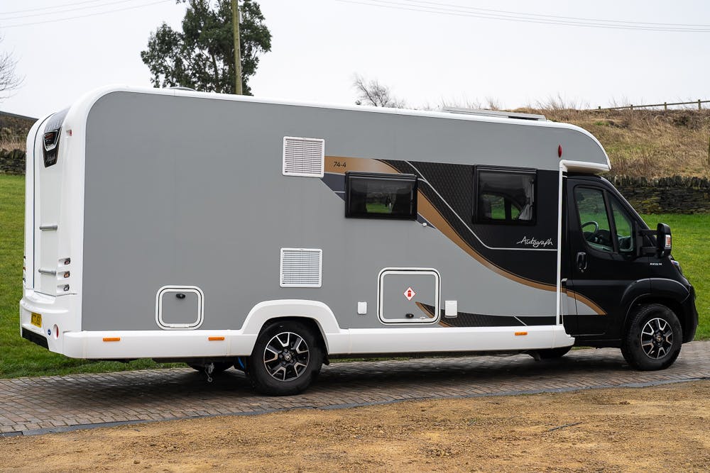 A side view of a gray and black 2022 Bailey Autograph 74-4 motorhome parked on a paved surface near a grassy area. The motorhome has a sleek design with various windows and vents visible, along with a door near the front. A tree and a wooden fence are visible in the background.