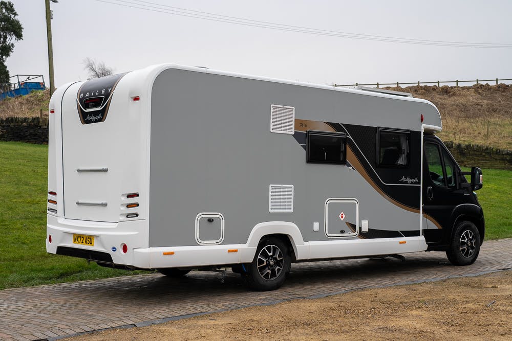 A large 2022 Bailey Autograph 74-4 recreational vehicle parked on a paved surface. The vehicle has a gray and black exterior with modern design elements, multiple windows, and a UK license plate. A grassy area and a fence are visible in the background.