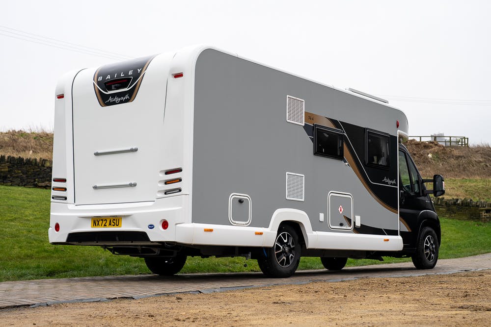 A 2022 Bailey Autograph 74-4 recreational vehicle (RV) with a sleek grey and white exterior is parked on a paved surface. The RV boasts several windows, a rear entrance, and ample exterior storage compartments. A grassy area and cloudy sky serve as the backdrop, completing the serene scene.