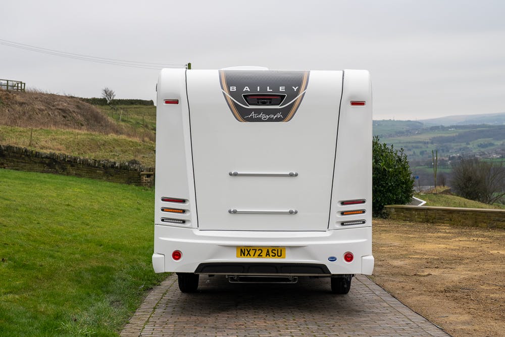 The image shows the rear view of a white 2022 Bailey Autograph 74-4 motorhome. The motorhome is parked on a paved surface with a grass area to the left and countryside scenery in the background. The license plate reads "NX72 ASU".