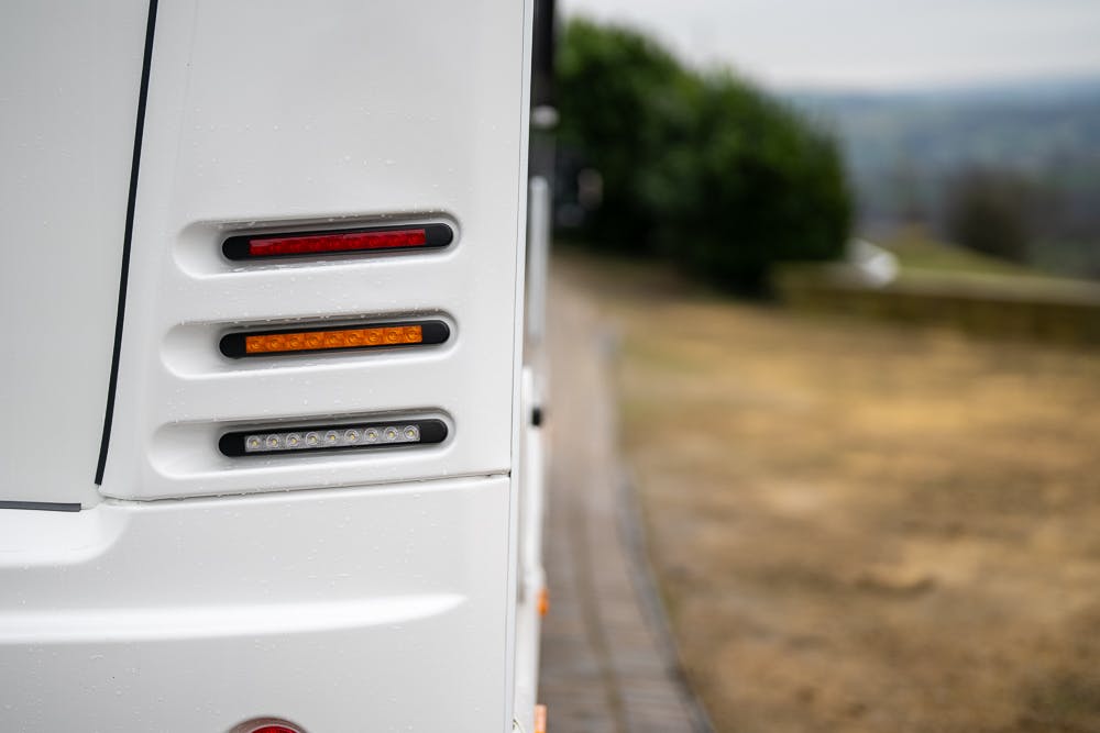 Close-up of the rear lights on the side of a 2022 Bailey Autograph 74-4. The lights include red, amber, and white sections. The background shows an out-of-focus view of a paved pathway and greenery.
