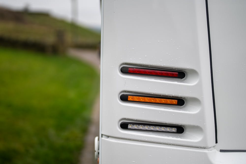 Close-up of the rear lights of a white 2022 Bailey Autograph 74-4, featuring three horizontal lights stacked vertically: a red light at the top, an amber light in the middle, and a clear light at the bottom. The background includes a blurred pathway and green grass.