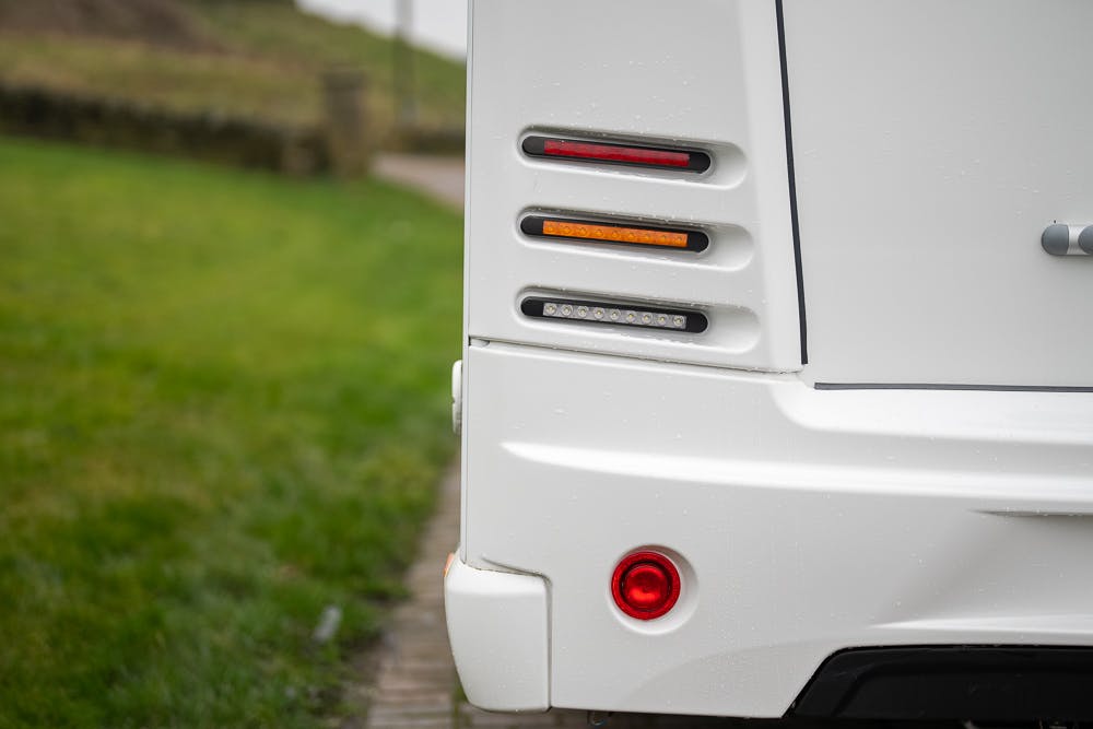 Close-up of the rear corner of a 2022 Bailey Autograph 74-4, showcasing its tail lights that include red, amber, and white lights. The vehicle is parked on a path next to a grassy area.