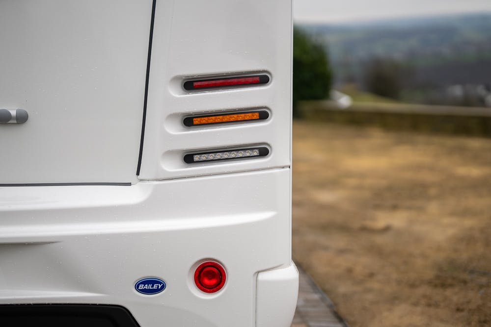 Close-up of the rear left side of a white 2022 Bailey Autograph 74-4 camper van featuring tail lights and brake lights. The van has a "Bailey" logo near the bottom and is parked on a gravel surface with a blurred, natural background.
