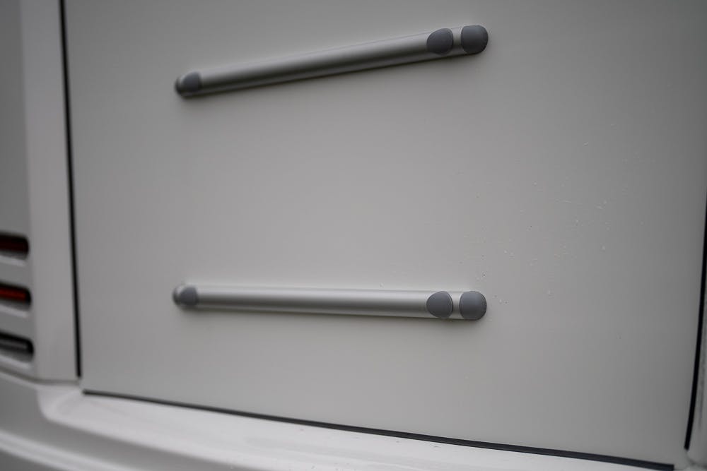 Close-up of a white surface featuring two horizontal metal rods attached with grey end caps on the 2022 Bailey Autograph 74-4. The background shows a faint outline of the structure’s form and edges, with part of a vehicle's backlight visible on the left side.