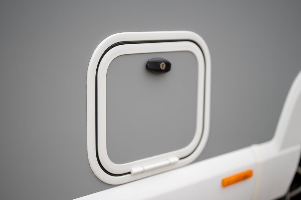 Close-up of a square, latched access door on the exterior of a gray 2022 Bailey Autograph 74-4, likely an RV or camper. The door has a black lock in the center and is framed with white trim. There is an orange reflector light visible to the bottom right of the door.