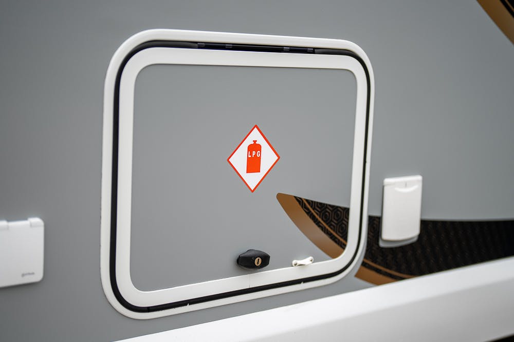 Close-up of a grey compartment door on a 2022 Bailey Autograph 74-4. The door features a red label with an image of a gas cylinder and the letters "LPG" indicating liquefied petroleum gas storage. There is a lock mechanism at the bottom of the door.