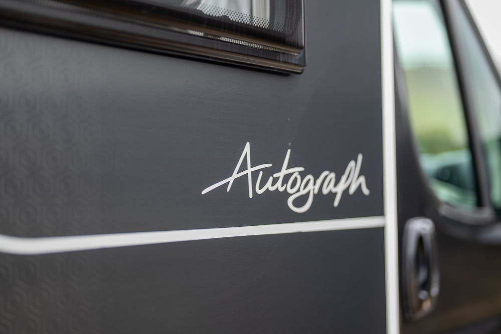 Close-up of the side of a 2022 Bailey Autograph 74-4, featuring a section of a window, part of a door with a handle, and the word "Autograph" written in a cursive font on the body. The vehicle's dark-colored exterior is accented with a thin white stripe.