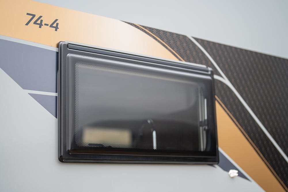Close-up of a rectangular black window with a partially transparent cover on the side of a 2022 Bailey Autograph 74-4. The side panel is grey with orange and black geometric designs. The number "74-4" is printed in the top left corner.