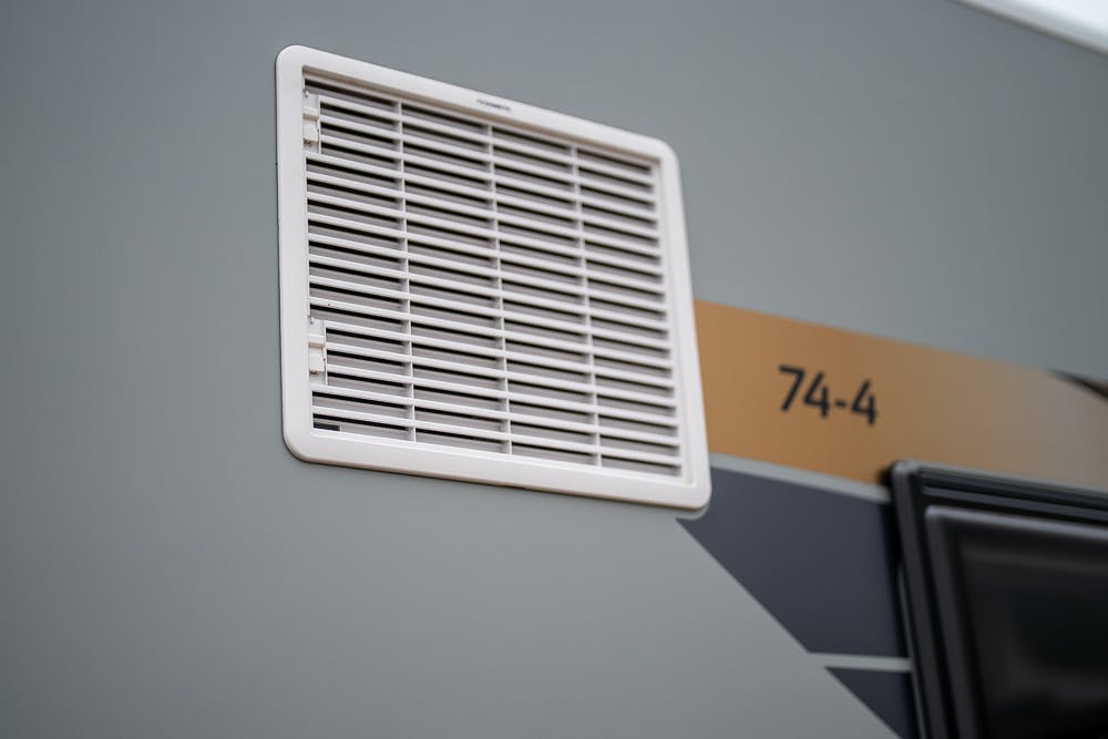Close-up of a white plastic vent attached to the exterior of a gray 2022 Bailey Autograph 74-4 vehicle, with the number 74-4 printed on it. The photo shows part of the window frame on the right side of the image.