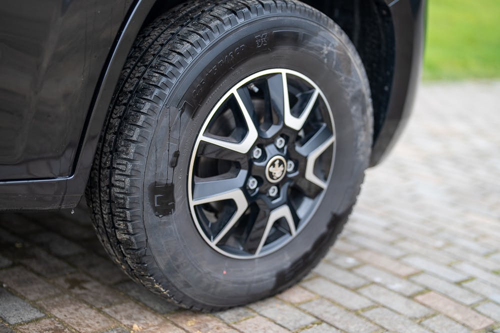 Close-up of the front left tire of a dark-colored 2022 Bailey Autograph 74-4. The tire is mounted on an alloy wheel with five double spokes, featuring a central logo. The vehicle is parked on a paved surface with grass visible in the background.