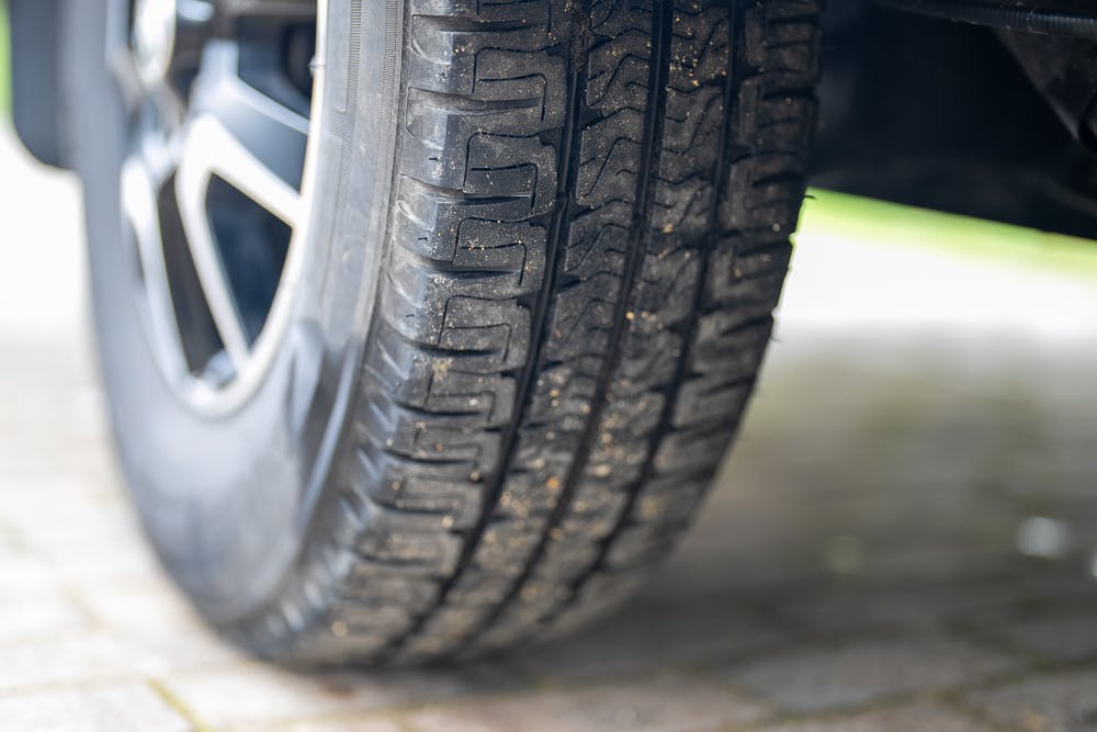 Close-up image of a car tire on a paved surface. The tire tread pattern is clearly visible, showing minor wear and traces of dirt. The car's wheel and part of the fender are also partially visible in the background, revealing a glimpse of the 2022 Bailey Autograph 74-4.