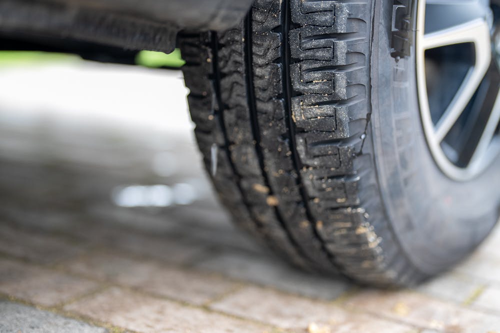 Close-up image of a car tire on a cobblestone surface. The tread pattern is clearly visible, and there is some dirt lodged in the grooves of the tire. Part of the alloy wheel from the 2022 Bailey Autograph 74-4 motorhome is also visible in the background.