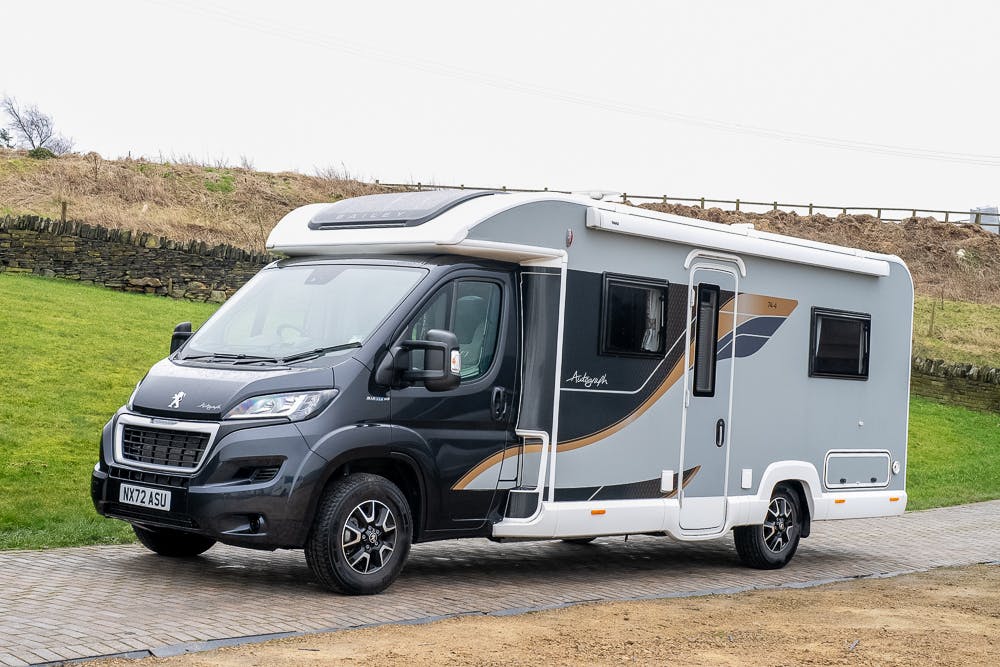 A gray and white 2022 Bailey Autograph 74-4 motorhome is parked on a paved surface with grass and a stone wall in the background. The vehicle's windows are tinted, featuring a metallic accent design on the side. The registration plate reads "NX17 AKU.