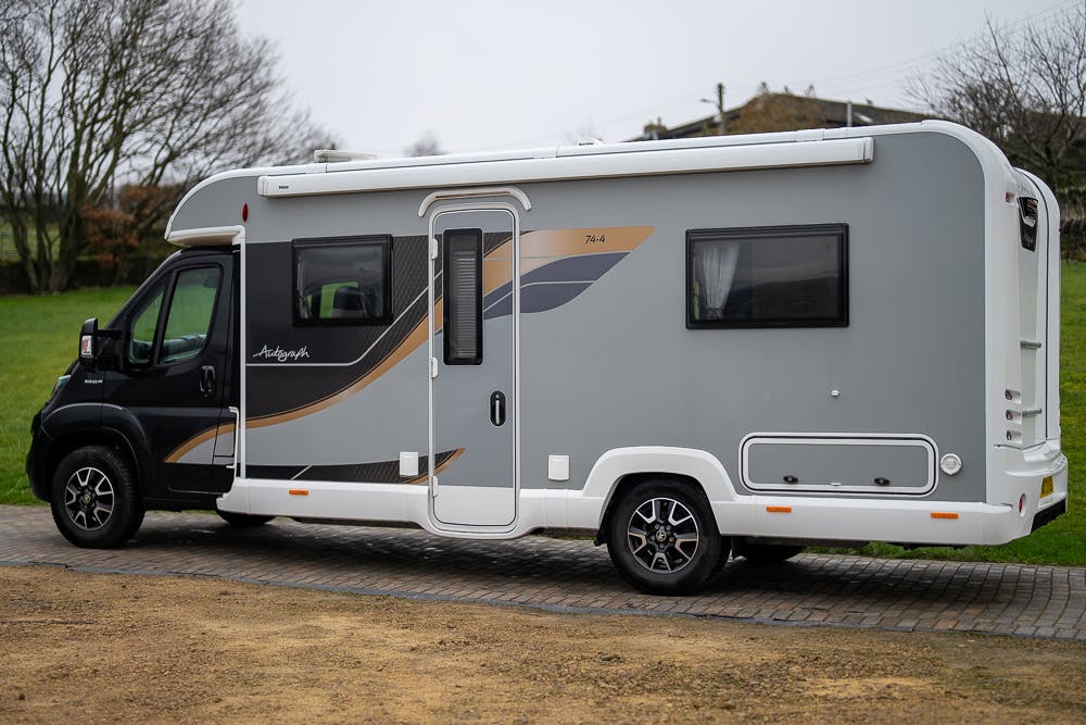 A modern, gray and white 2022 Bailey Autograph 74-4 motorhome parked on a cobbled path with a grassy area and trees in the background. The motorhome features large windows and geometric decals on the side, along with alloy wheels and a built-in door step.