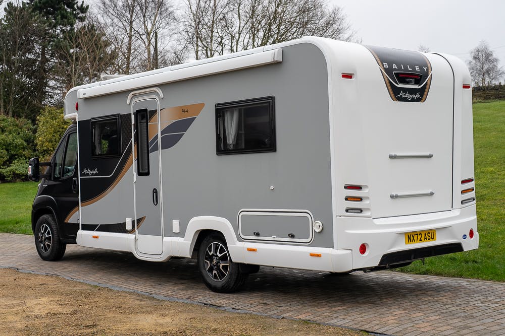 A 2022 Bailey Autograph 74-4 motorhome is parked on a paved driveway. It features a grey and white exterior with stylized accents and a rear window. The motorhome's tail lights, side door, and license plate "NX72 ASU" are visible. Trees are in the background.