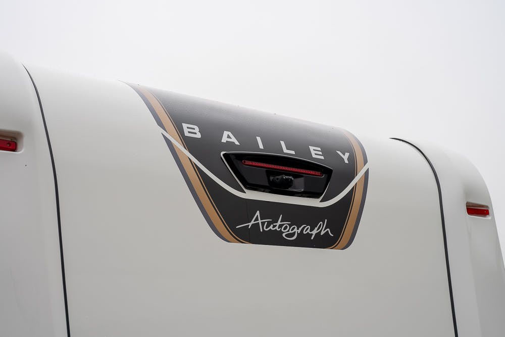 Close-up of the rear section of a 2022 Bailey Autograph 74-4 motorhome, featuring the brand name "BAILEY" at the top and the "Autograph" label below, set against a black and gold accent design. The image focuses on the upper portion of this stylish vehicle.
