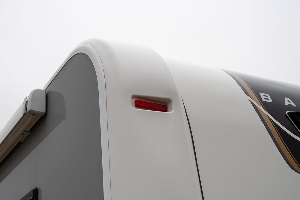 Close-up of the rear upper corner of a 2022 Bailey Autograph 74-4. The image shows a white and gray exterior with a small red light near the top edge. The RV's side has a curved design element, and the sky in the background is overcast.