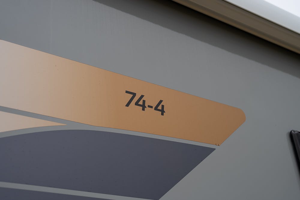 A close-up of a section on an RV exterior displaying the marking "74-4" in black text on a beige background. The image focuses on a specific part of the 2022 Bailey Autograph 74-4's side panel, showcasing the detail of its distinctive graphic design.