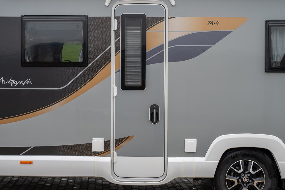 Close-up view of the central door of the 2022 Bailey Autograph 74-4. The design of the RV's exterior showcases a combination of grey, black, and beige colors with accent lines and a decorative pattern.