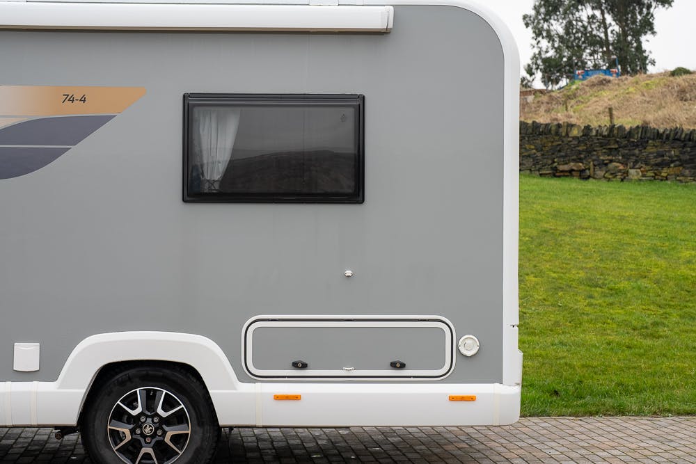 A close-up view showing the rear side of a gray 2022 Bailey Autograph 74-4 camper van with a small window and storage compartment. A portion of a tire and wheel is visible on the lower left. The background includes grass, a stone wall, and a cloudy sky.