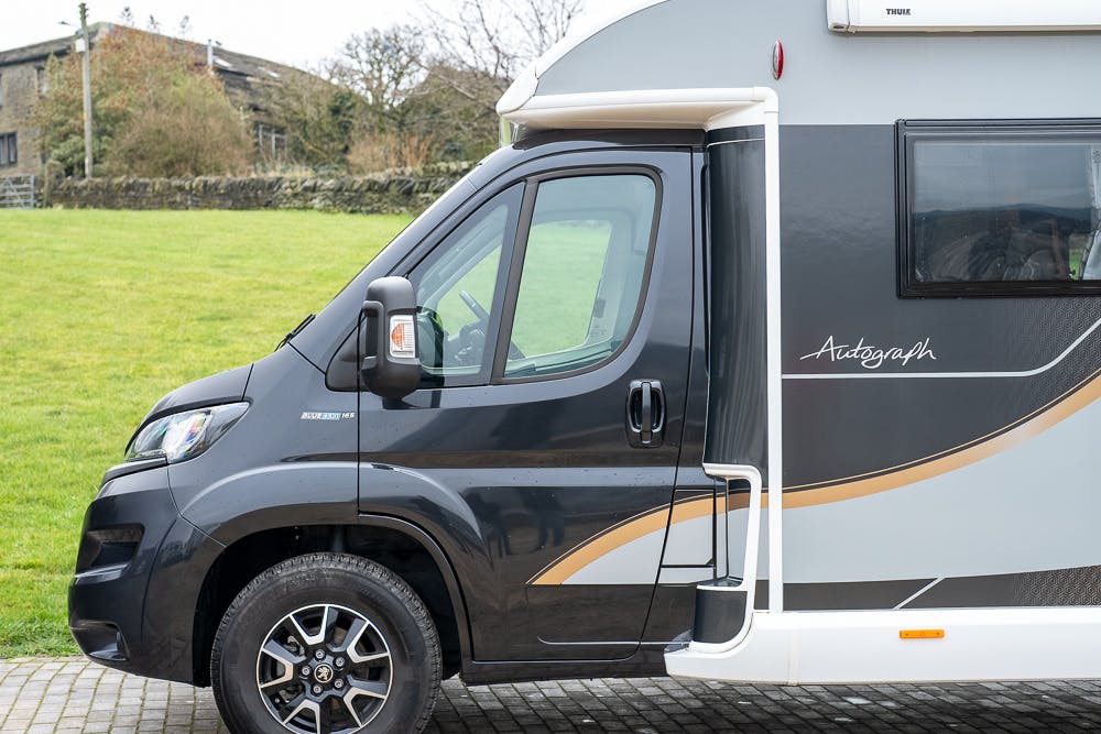 A partial side view of a black and silver motorhome with the word "Autograph" written on the side. The 2022 Bailey Autograph 74-4 is parked on a paved area with a grassy field and stone wall in the background.
