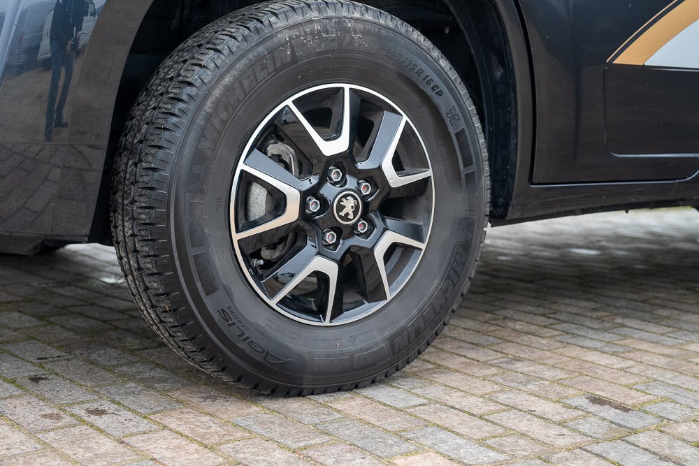 Close-up of a black 2022 Bailey Autograph 74-4's front wheel, showing a five-spoke alloy rim and a Michelin tire. The motorhome is parked on a cobblestone surface, and the surrounding area is partially visible, reflecting off the vehicle's dark exterior.