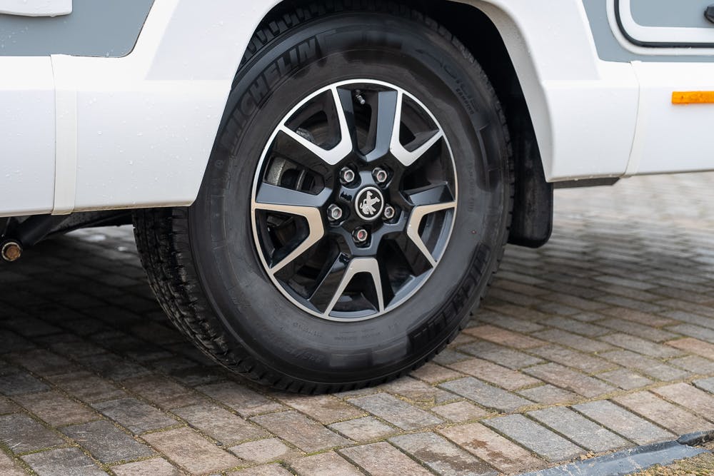 A close-up view of a 2022 Bailey Autograph 74-4’s wheel and tire, featuring a black and silver alloy rim and Michelin tire. The vehicle is parked on a paved surface made of small, rectangular bricks. The lower body of the vehicle is white and gray.