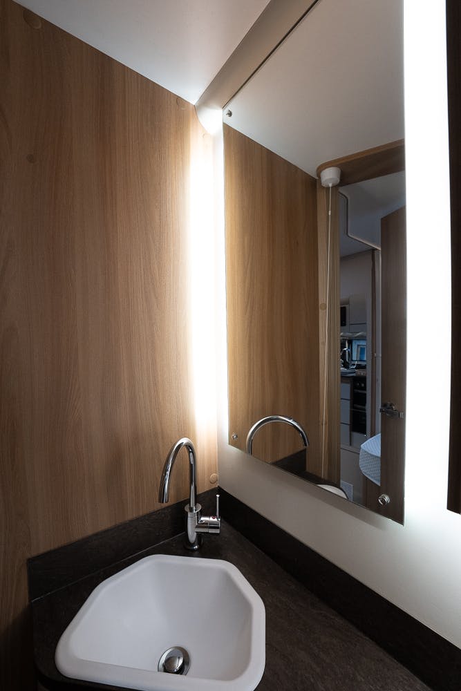 A bathroom sink with a modern design is shown in the 2022 Bailey Autograph 74-4. The dark-colored countertop complements the chrome faucet, and a large mirror with vertical lighting on the left side is mounted on the wooden-paneled wall. A partial view of the adjoining room is visible.