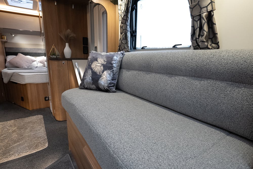 The image shows the interior of a modern 2022 Bailey Autograph 74-4 RV. There is a grey upholstered sofa with a decorative pillow featuring a feather design. In the background, there is a glimpse of a bedroom with a bed, nightstand, and small wooden shelf. The floor is carpeted.