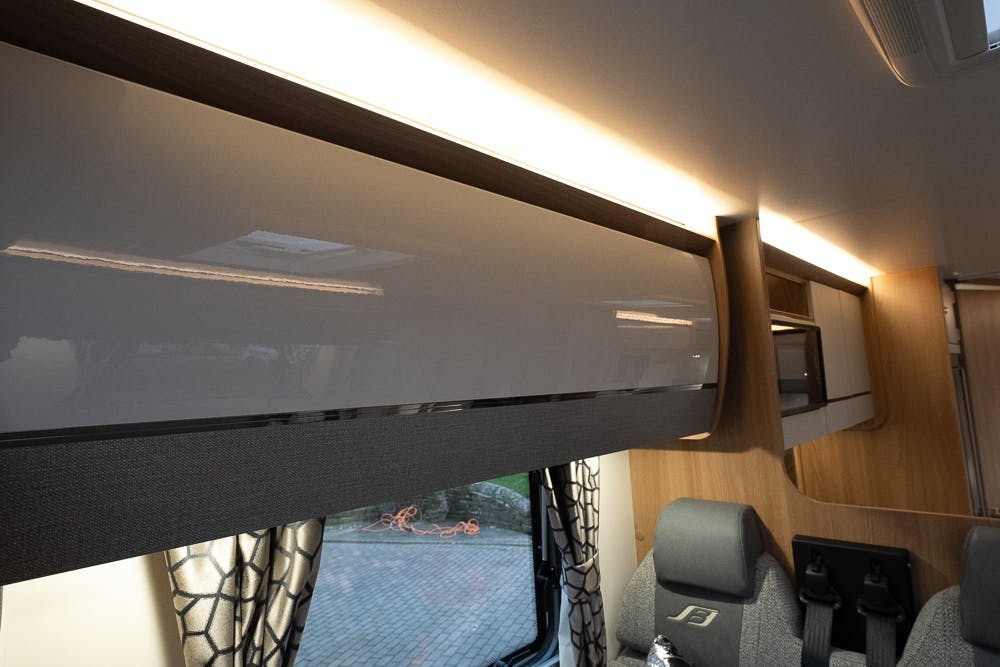 The image shows the interior of a 2022 Bailey Autograph 74-4 motorhome. A row of modern, glossy storage cabinets runs along the top of the wall. Below, there is a window with patterned curtains. Two upholstered seats are partially visible at the bottom right.