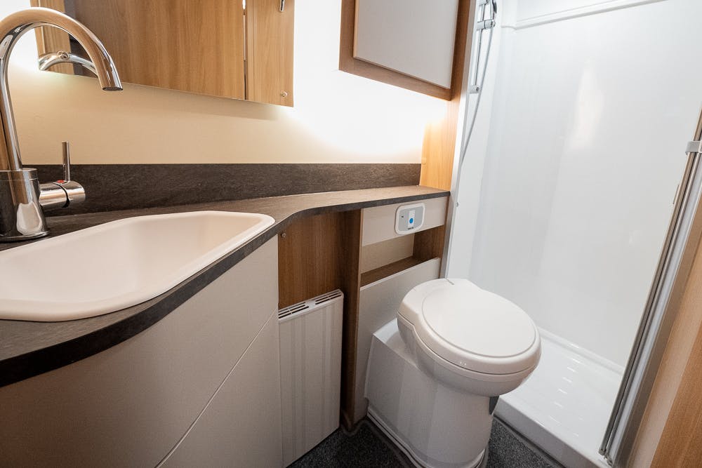 A compact bathroom in the 2022 Bailey Autograph 74-4 features a white sink with a curved counter, a chrome faucet, a mirror cabinet, a white toilet, and a shower enclosure with a glass door. The countertop and cabinetry are wooden with a dark laminate finish.